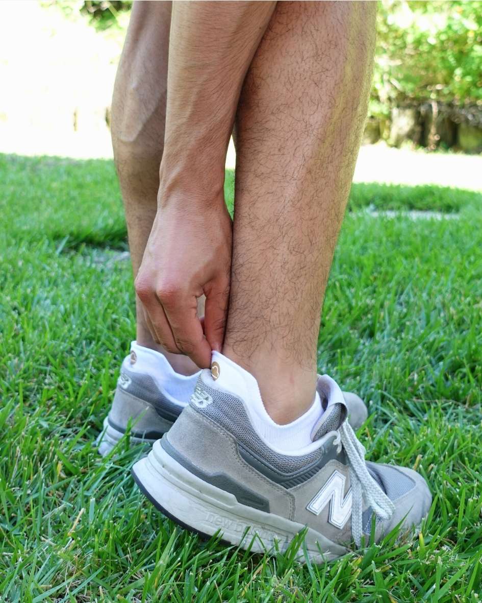 Person outdoors on grass pulling on a white padded ankle sock beside a white sneaker