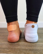 Two feet side by side, one bare heel with visible redness and one wearing a white padded ankle sock in a sneaker